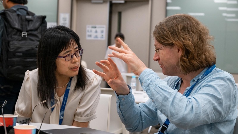 Prof. Defang Duan and Prof. Chris Pickard sit at a table in a conference setting, engaged in a focused discussion. Both wear lanyards, and a laptop, cups, and posters in the background indicate an academic event.