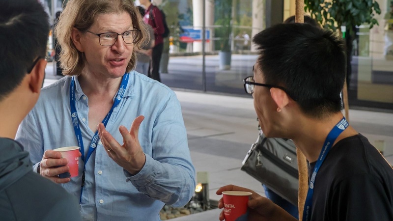 Three conference attendees stand talking in a modern lobby area. The man in the centre, wearing glasses and a light blue shirt, gestures while holding a red paper cup. Another attendee holding a similar cup listens beside him, and a third person is visible from behind. Blue lanyards and the busy background suggest an academic event.