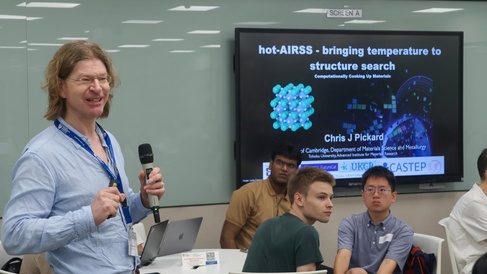 “Prof. Chris J. Pickard stands speaking into a microphone at a symposium at Nanyang Technological University. He faces an audience seated in a lecture room. Behind him, a large presentation slide titled ‘hot‑AIRSS – bringing temperature to structure search’ is displayed, featuring molecular graphics and logos for UKCP, CASTEP, and FutureCat