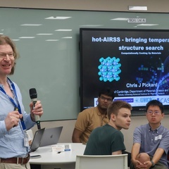 “Prof. Chris J. Pickard stands speaking into a microphone at a symposium at Nanyang Technological University. He faces an audience seated in a lecture room. Behind him, a large presentation slide titled ‘hot‑AIRSS – bringing temperature to structure search’ is displayed, featuring molecular graphics and logos for UKCP, CASTEP, and FutureCat